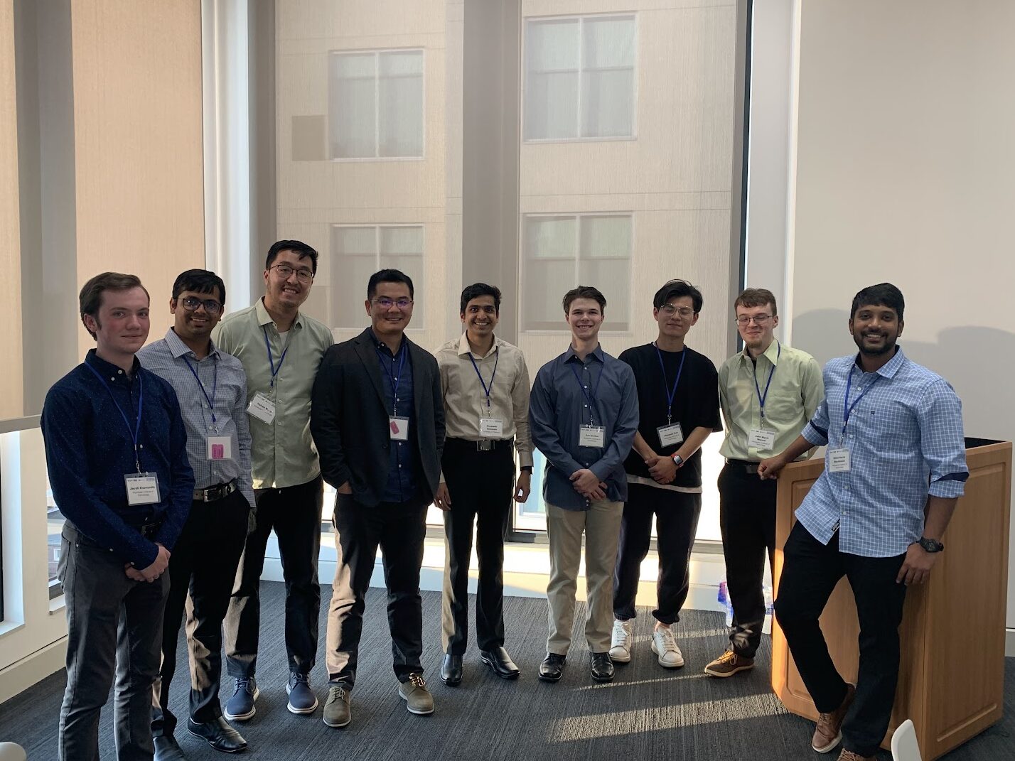 Nine conference attendees wearing name badges stand together in a bright room near large windows and a presentation podium. The group photo appears to document participation in the 10th Graphene and Beyond Workshop at Penn State University.