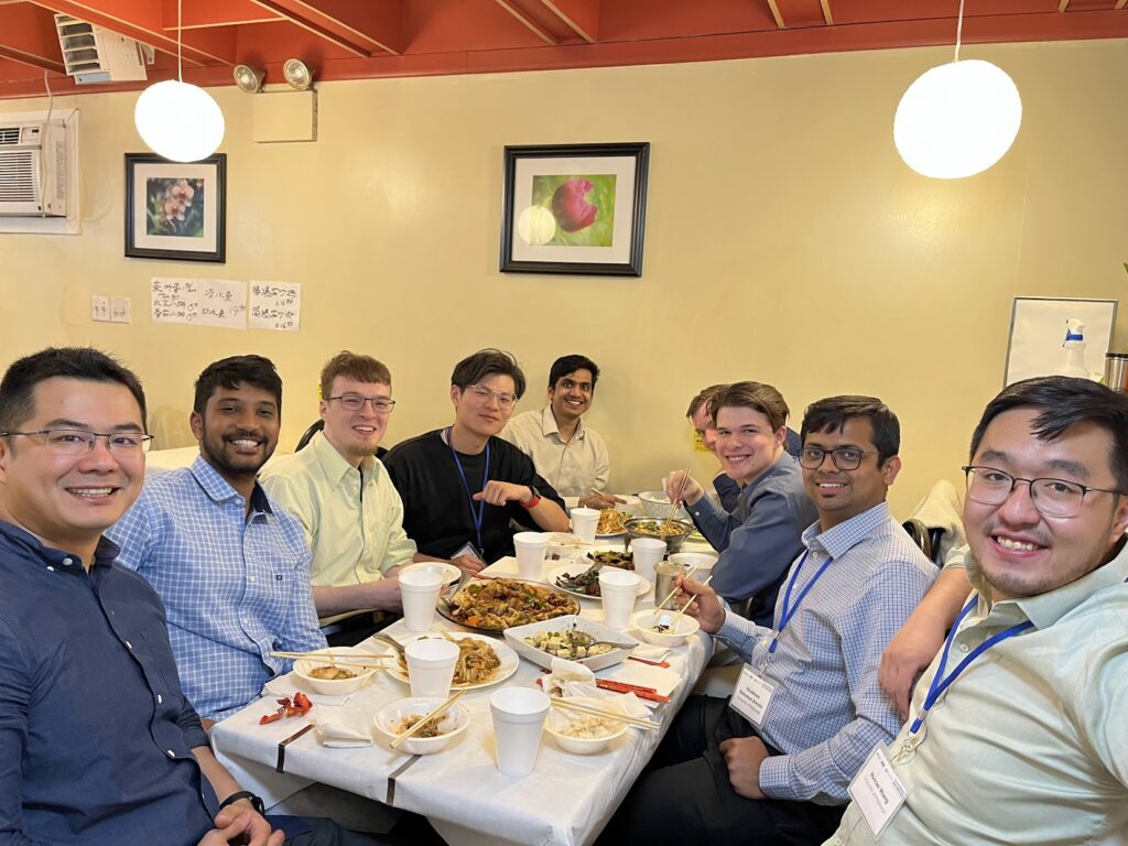 A group of conference attendees sit around a restaurant table with shared dishes, bowls, cups, and chopsticks after the workshop. The dining area has framed wall art, hanging lights, and a casual group-meal setting.