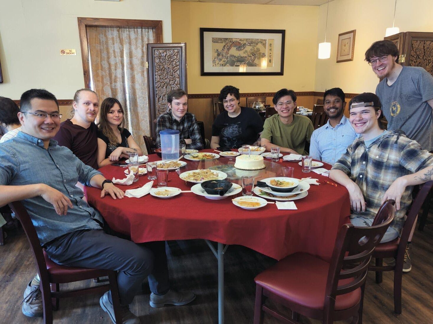 Members of the Xu group sit around a large restaurant table with plates, glasses, serving dishes, and a cake while celebrating the end of a successful semester. The dining room includes a red tablecloth, framed wall art, pendant lights, and wood-paneled décor.