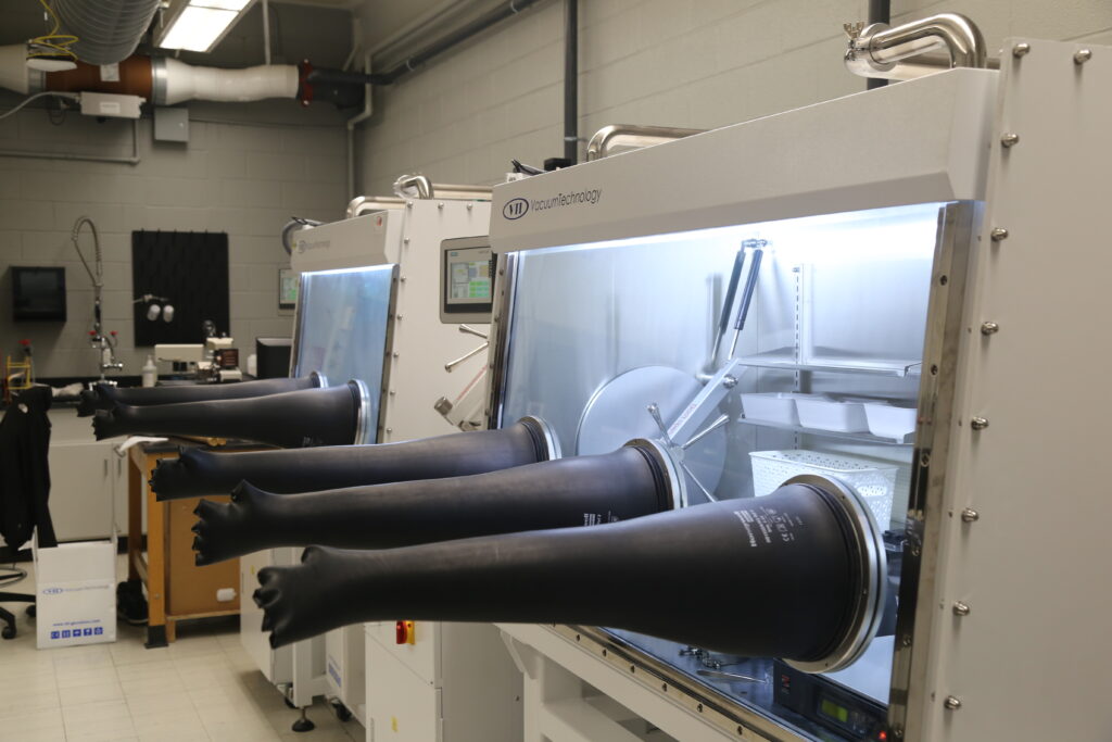 A newly installed 2+3 bay glovebox fills the foreground of a laboratory, with several black glove ports extending from illuminated enclosed work chambers. Additional lab benches, ventilation ducts, sinks, and equipment are visible in the background.