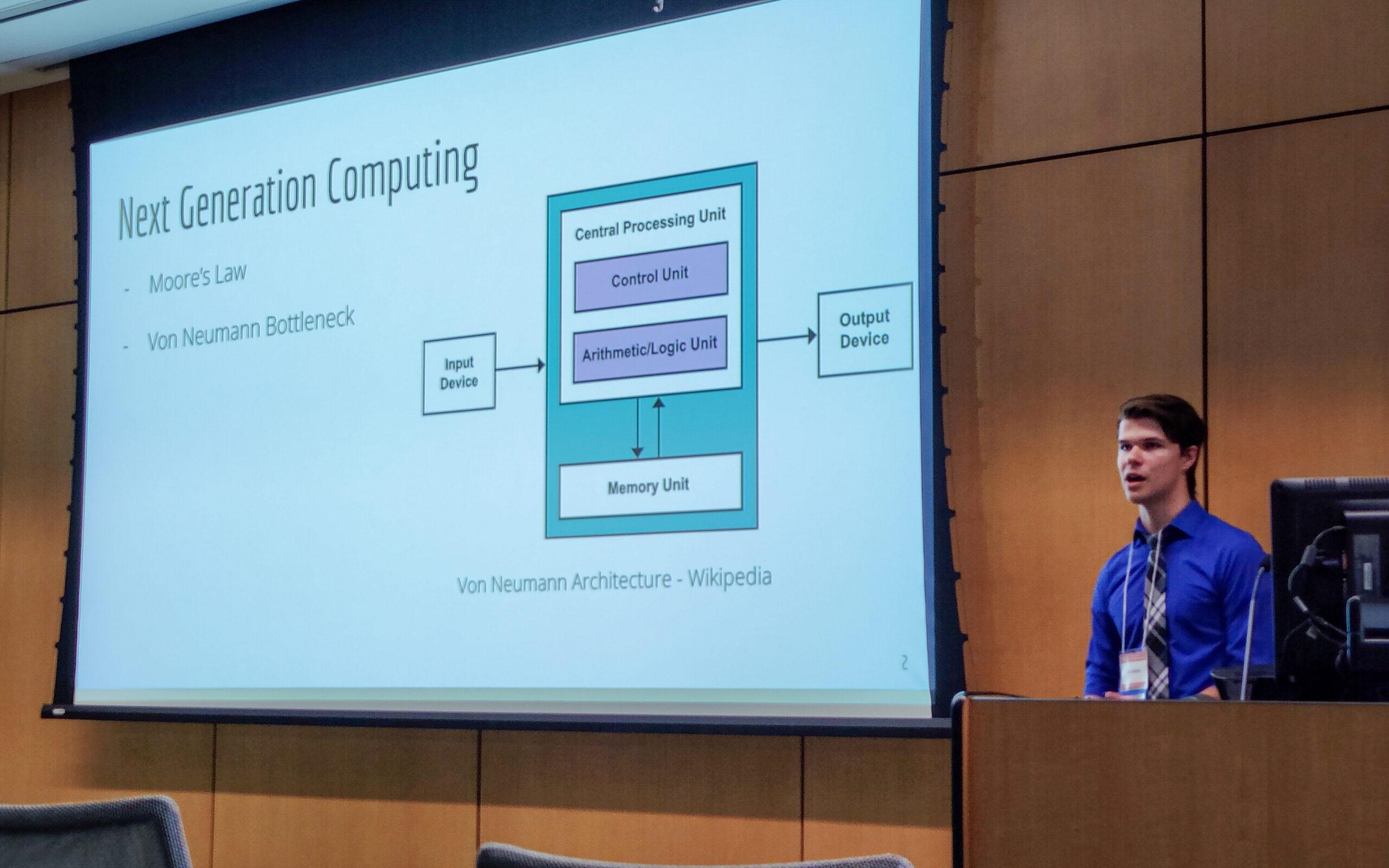 A presenter stands beside a large projected slide titled “Next Generation Computing,” which includes bullet points about Moore’s Law and the Von Neumann bottleneck. The slide also shows a diagram of a central processing unit connected to input, output, and memory units.