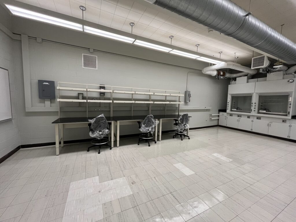 Clean, empty laboratory space with newly installed lab benches, shelving, wrapped chairs, overhead lighting, exposed ventilation ducts, and fume hoods along the back wall. The room appears prepared for equipment setup and future research use.