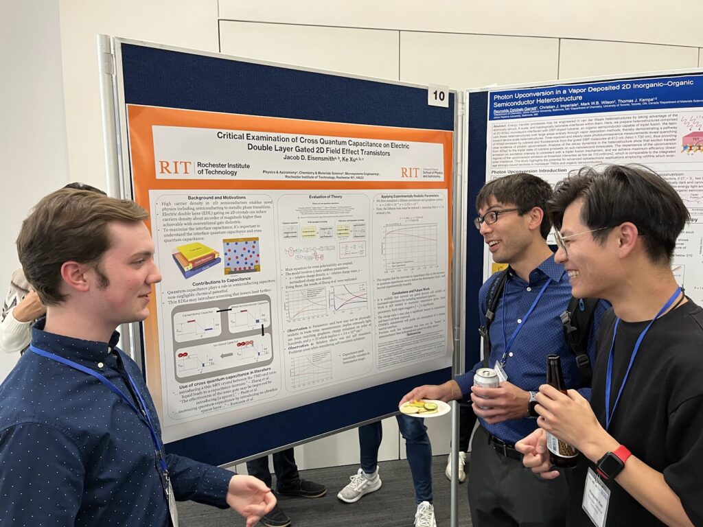 A presenter discusses an RIT research poster titled “Critical Examination of Cross Quantum Capacitance on Electric Double Layer Gated 2D Field Effect Transistors” with two attendees. The poster includes device schematics, equations, and graphs, with another poster displayed on the adjacent board.