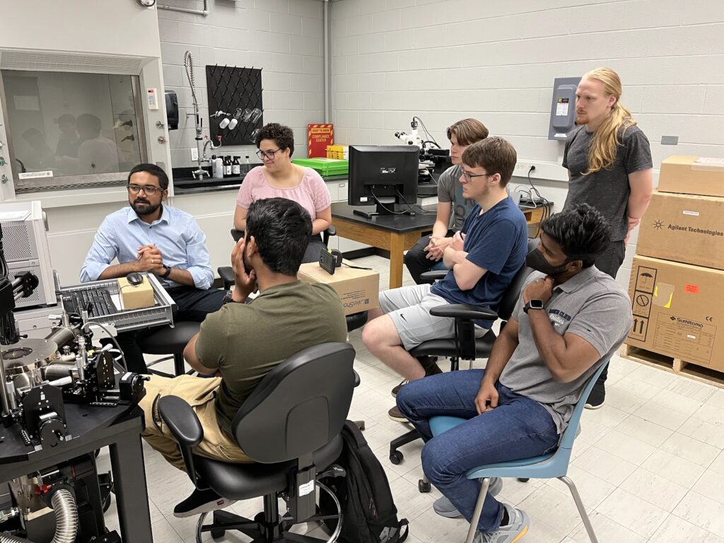 Several people sit and stand in a lab around a semiconductor probe station and computer equipment during training on a Keysight Semiconductor Parameter Analyzer. Lab benches, microscopes, fume hoods, boxes, and measurement equipment are visible around the room.