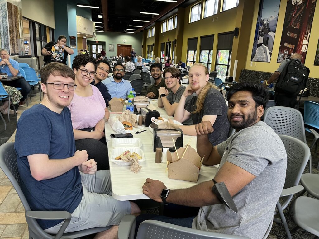 The training group sits together at a long table in a campus dining area with takeout containers, drinks, and bags on the table. Large windows, café seating, and other people are visible in the background.