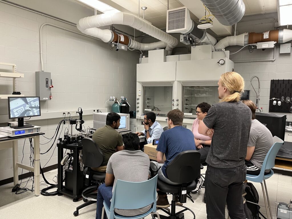 A group gathers closely around a probe station and computer in a lab while viewing semiconductor measurement equipment during training. A monitor displays a magnified device image, with fume hoods, ventilation ducts, cables, and lab instruments in the background.