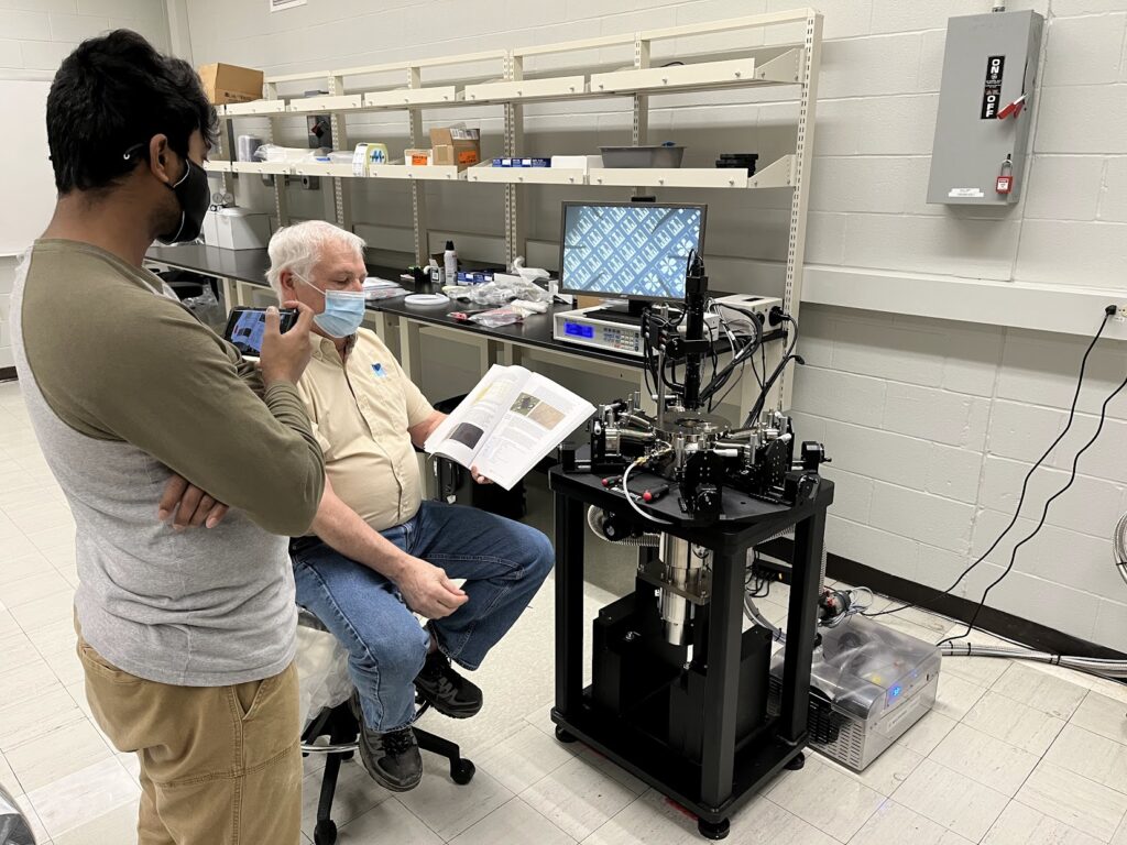 A person observes training on a newly installed Lake Shore Cryogenic Probe Station while another person seated nearby holds an open manual. The lab setup includes the probe station, microscope components, monitor, control equipment, shelving, and connected cables.