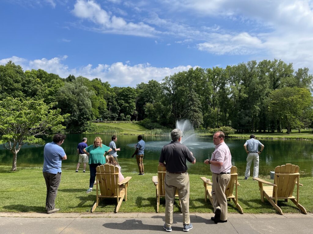Several people gather outdoors near a pond with wooden chairs, a fountain, and trees in the background during a SoPA faculty retreat. The scene shows small groups standing and talking on a sunny day beside the water.