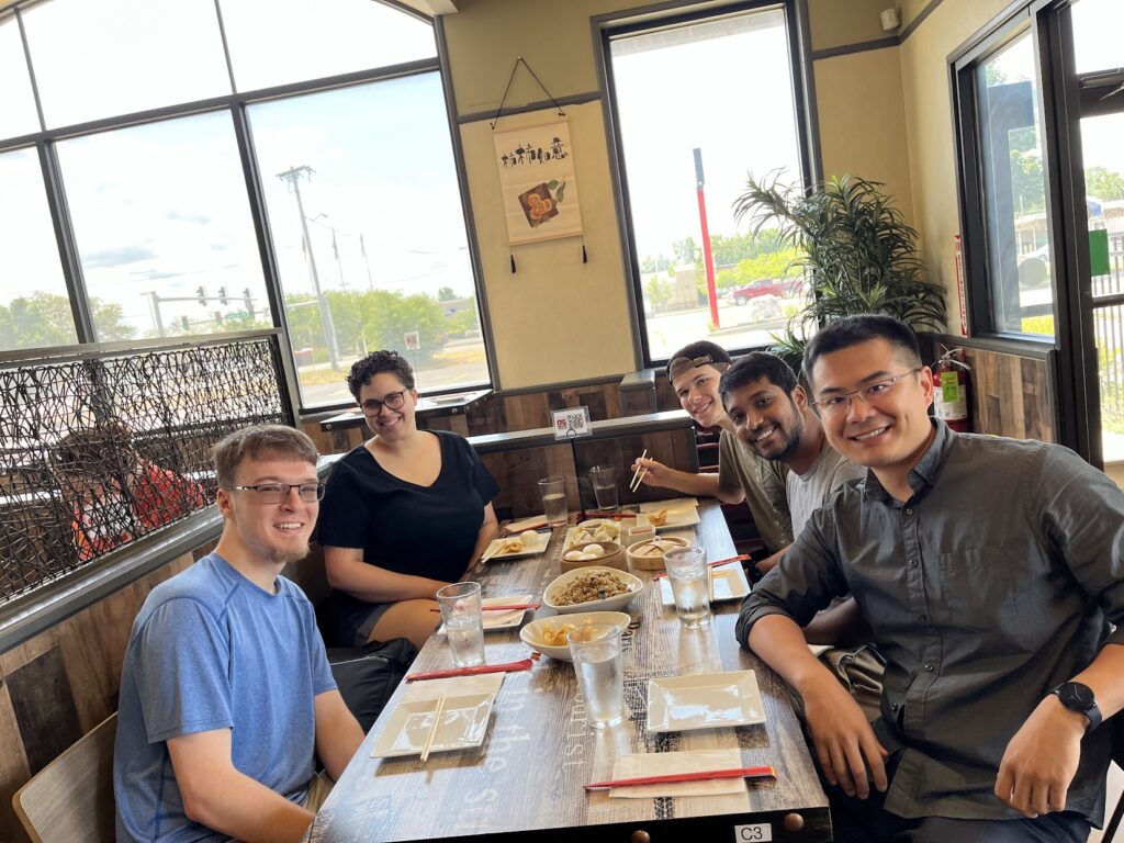 Five people sit around a restaurant table with plates of food, water glasses, chopsticks, and bamboo steamer baskets of xiaolongbao. Large windows, indoor plants, and wall decor are visible in the background during the group’s break from summer research.