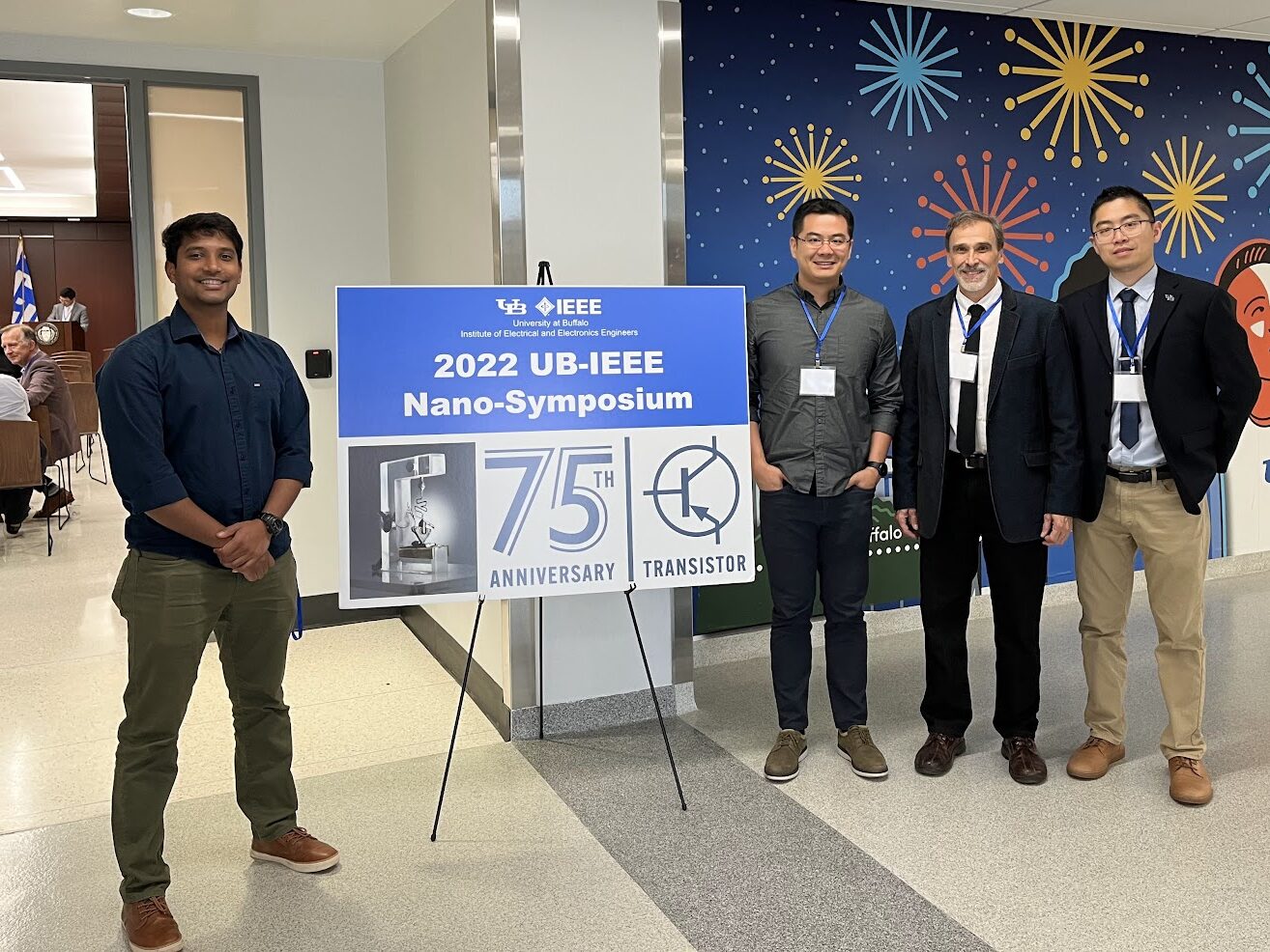 Four attendees stand beside a sign for the 2022 UB-IEEE Nano-Symposium at the University at Buffalo, which also marks the 75th anniversary of the transistor. A colorful wall mural and a seated audience area are visible in the background.