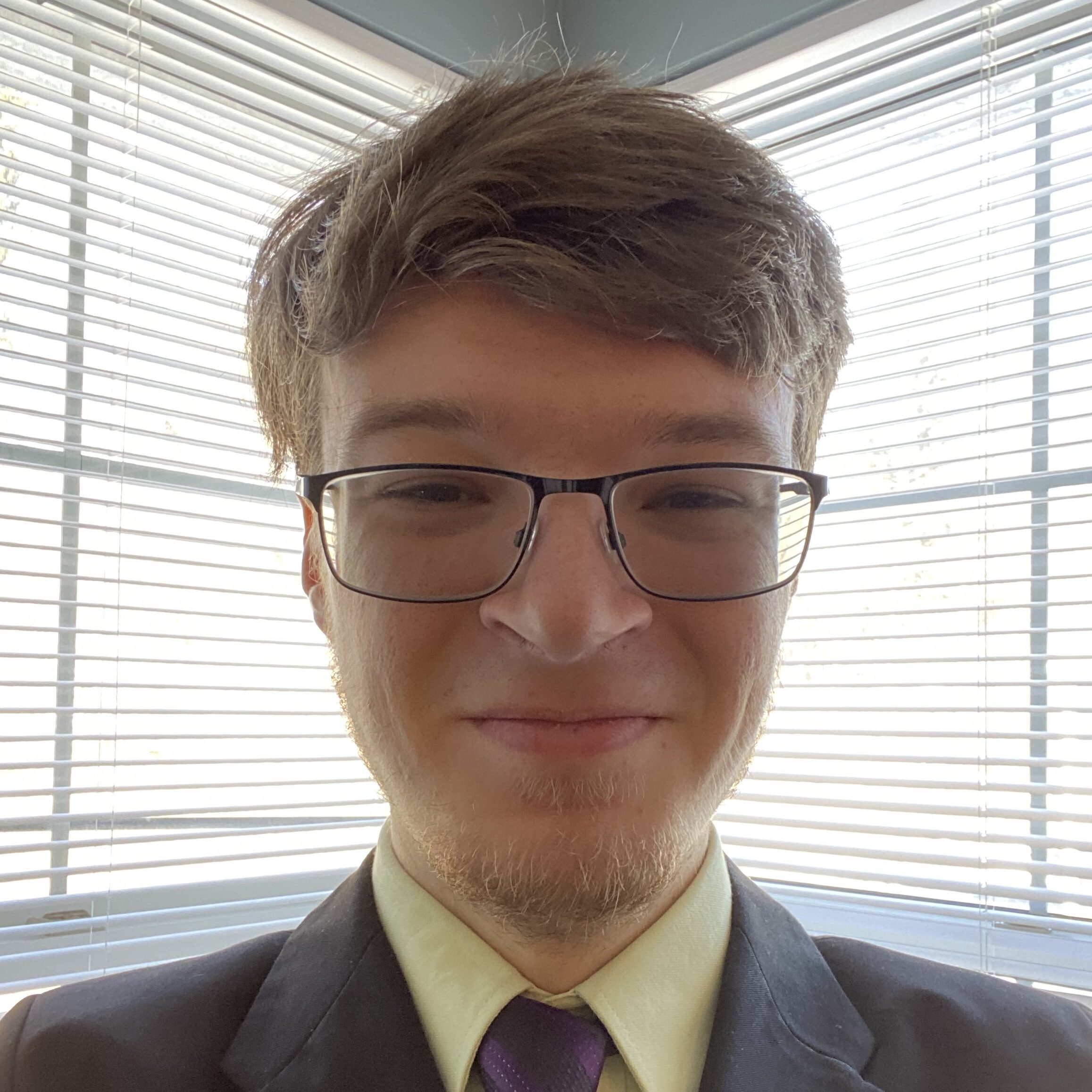 Head-and-shoulders portrait of a person wearing glasses, a suit jacket, collared shirt, and tie, seated in front of bright window blinds.