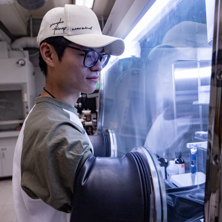 Huayuan Han working in a laboratory glovebox while wearing glasses and a baseball cap.