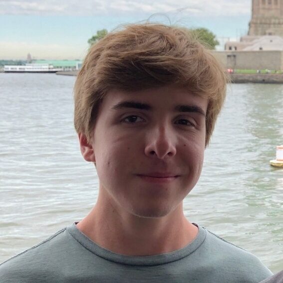 Gavin Heil standing near the water with a city landmark in the background, wearing a light green shirt.