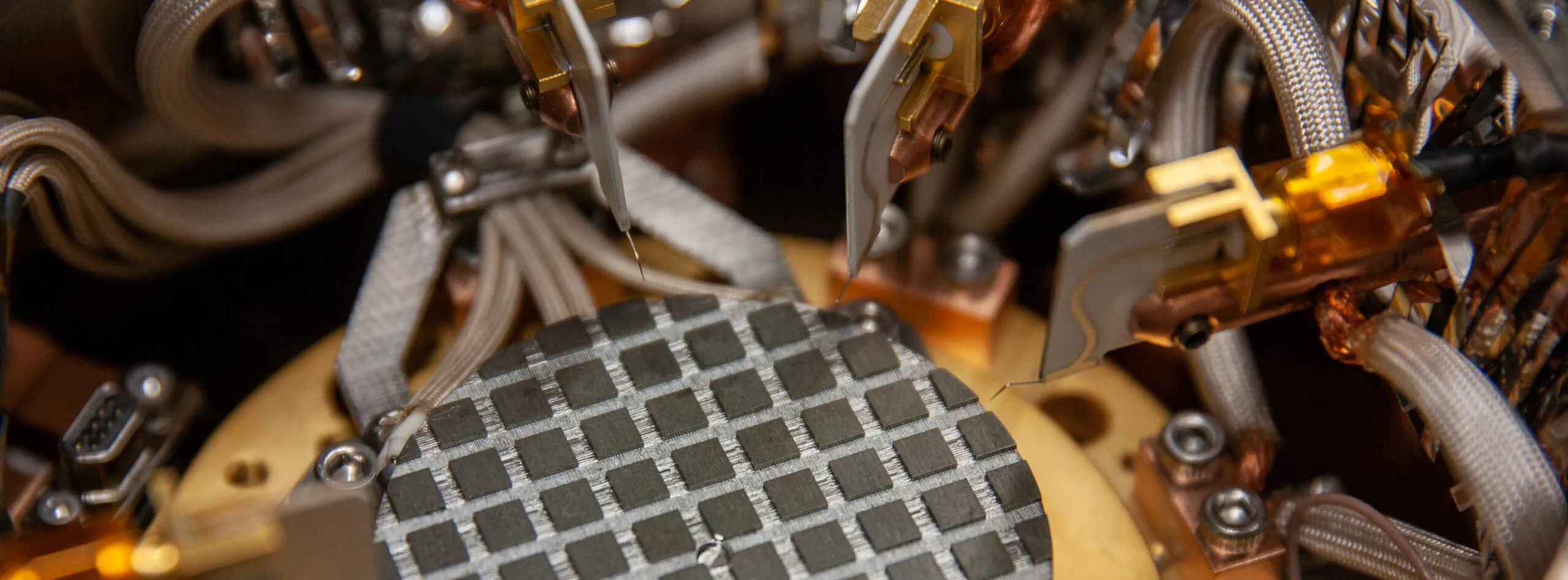 Close-up of a semiconductor wafer with a grid pattern, surrounded by precision probes and cables inside a testing or fabrication machine.