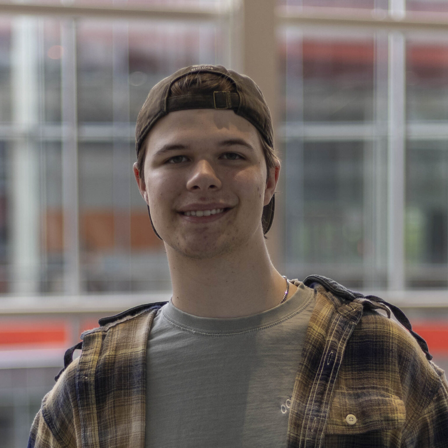 Cori Sutton smiling indoors in front of large windows, wearing a backward cap and plaid shirt.