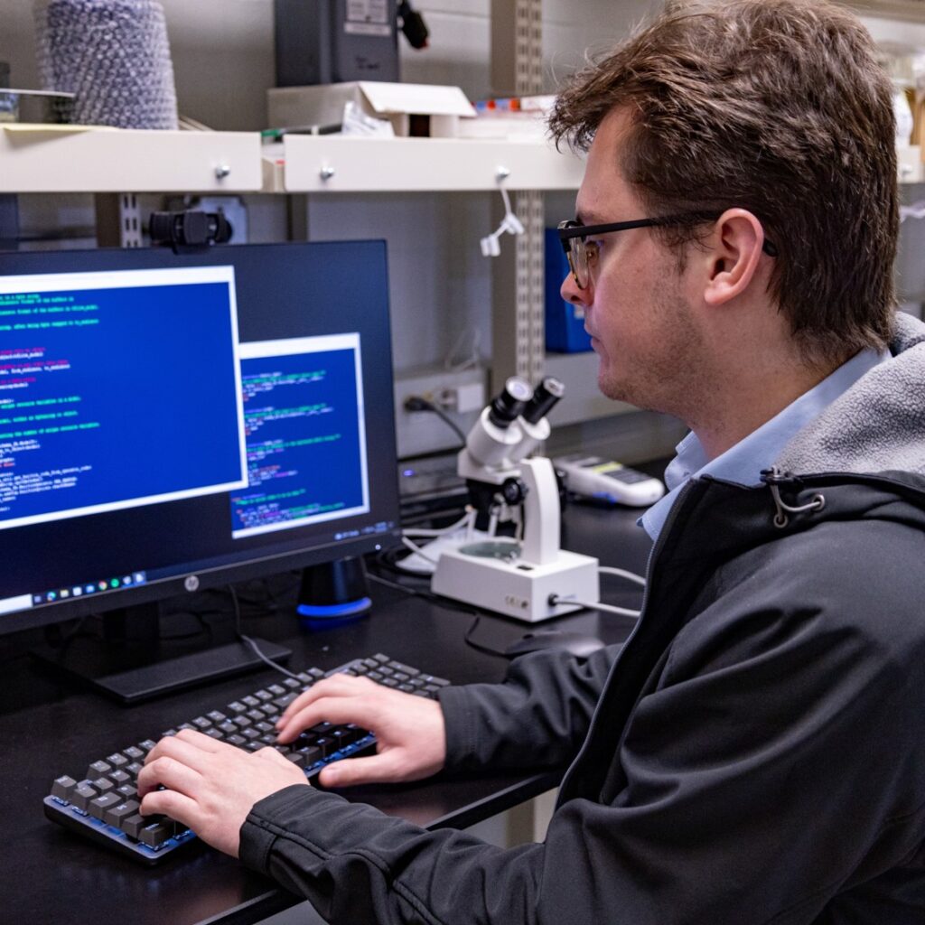Jake Streamer working at a computer in a lab, with code on the monitor and a microscope nearby.