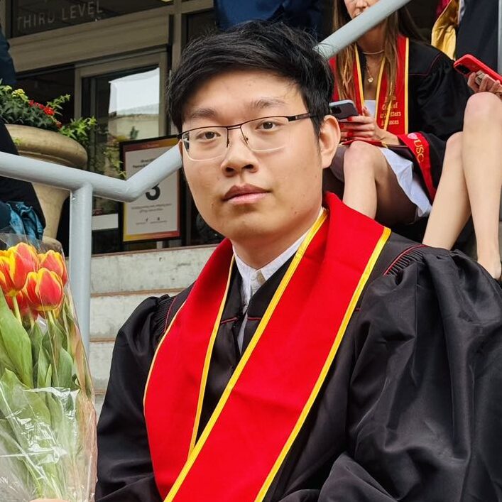 Jimmy Li seated outdoors in graduation attire, wearing a red stole and holding flowers.