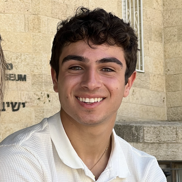 Joshua Masri smiling outdoors in front of a stone building, wearing a white collared shirt.