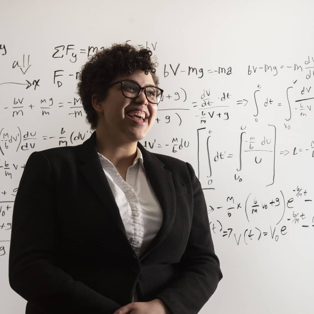 Claire Dantzlerward smiling in front of a whiteboard covered with physics equations, wearing a blazer and button-down shirt.